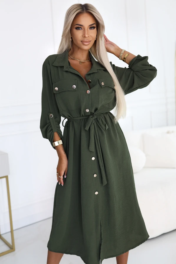 Stylowa sukienka koszulowa w kolorze khaki, z wiązaniem w pasie, guzikiem zapinana na całej długości, długi rękaw z podpięciem, elegancki fason marki Numoco Basic, europejska produkcja, prezentacja na modelce w nowoczesnym wnętrzu.