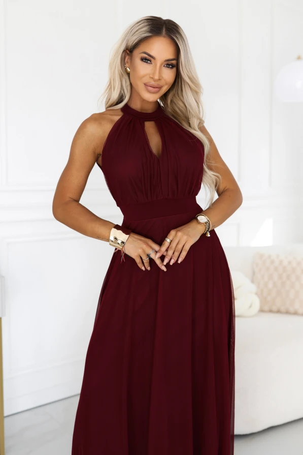 Modelka w bordowej sukience maxi z dekoltem halter, wycięciem w kształcie łezki i marszczoną górą. Szeroki pas podkreśla talię, a rozkloszowany dół z siatki ma podszewkę i transparentne wykończenie przy kostkach.