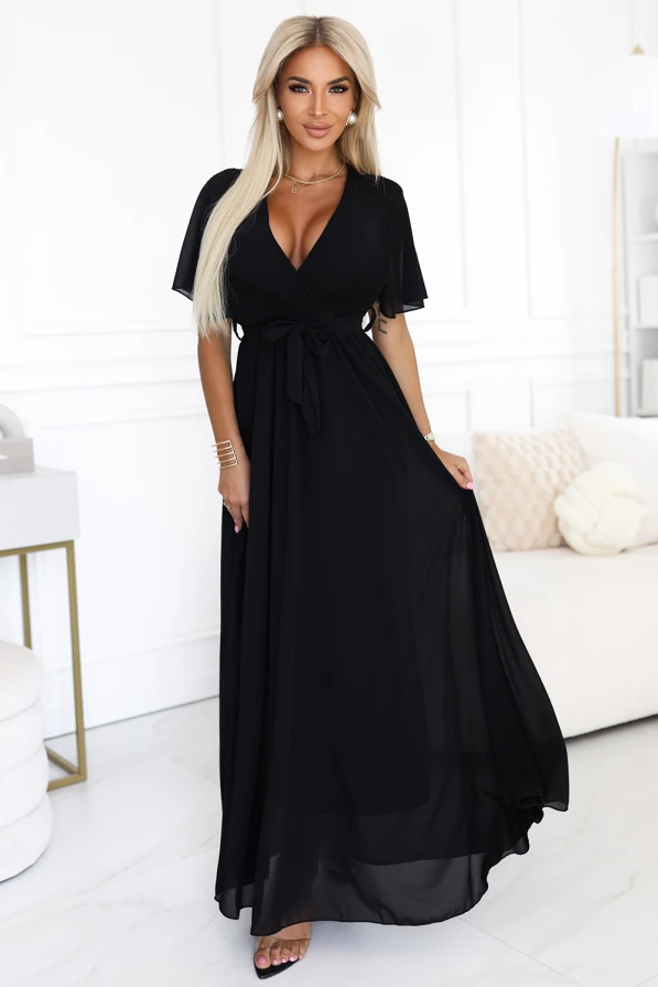 Modelka prezentuje elegancką czarną sukienkę maxi z szyfonu, z plisowanym dekoltem w kształcie litery V, krótkimi rękawkami i wiązaniem w talii; lekka i kobieca stylizacja marki Numoco Basic, idealna na wyjątkowe okazje.