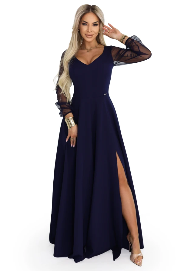 Modelka w eleganckiej, granatowej sukni maxi z dekoltem V, transparentnymi długimi rękawami i rozcięciem na nodze, prezentująca wieczorową stylizację na wesele, galę lub przyjęcie w jasnym, minimalistycznym wnętrzu.